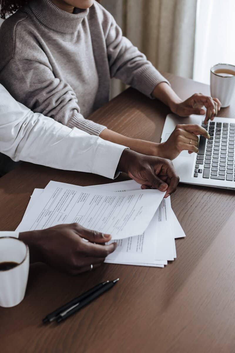 A person using a laptop next to someone reviewing paperwork.