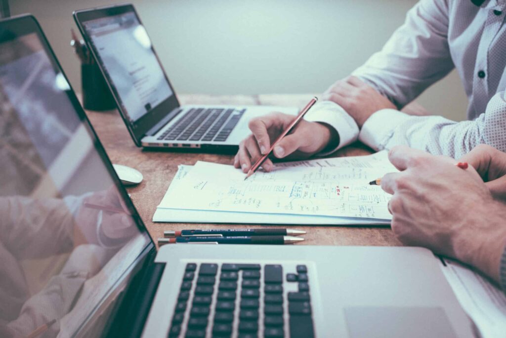 Photo shows hands of two people looking at paperwork over two open laptop computers